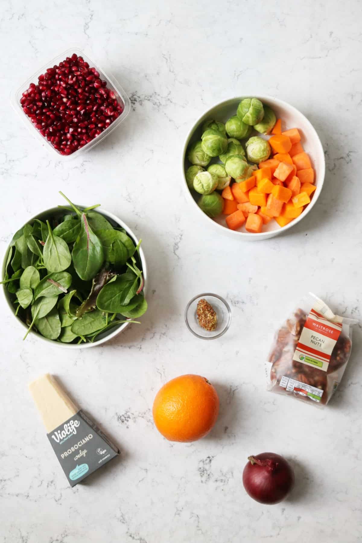 Pomegranate, Brussels sprouts, butternut squash, mixed salad leaves, wholegrain mustard, pecans, vegan Parmesan, orange and red onion laid out on a white marble counter top