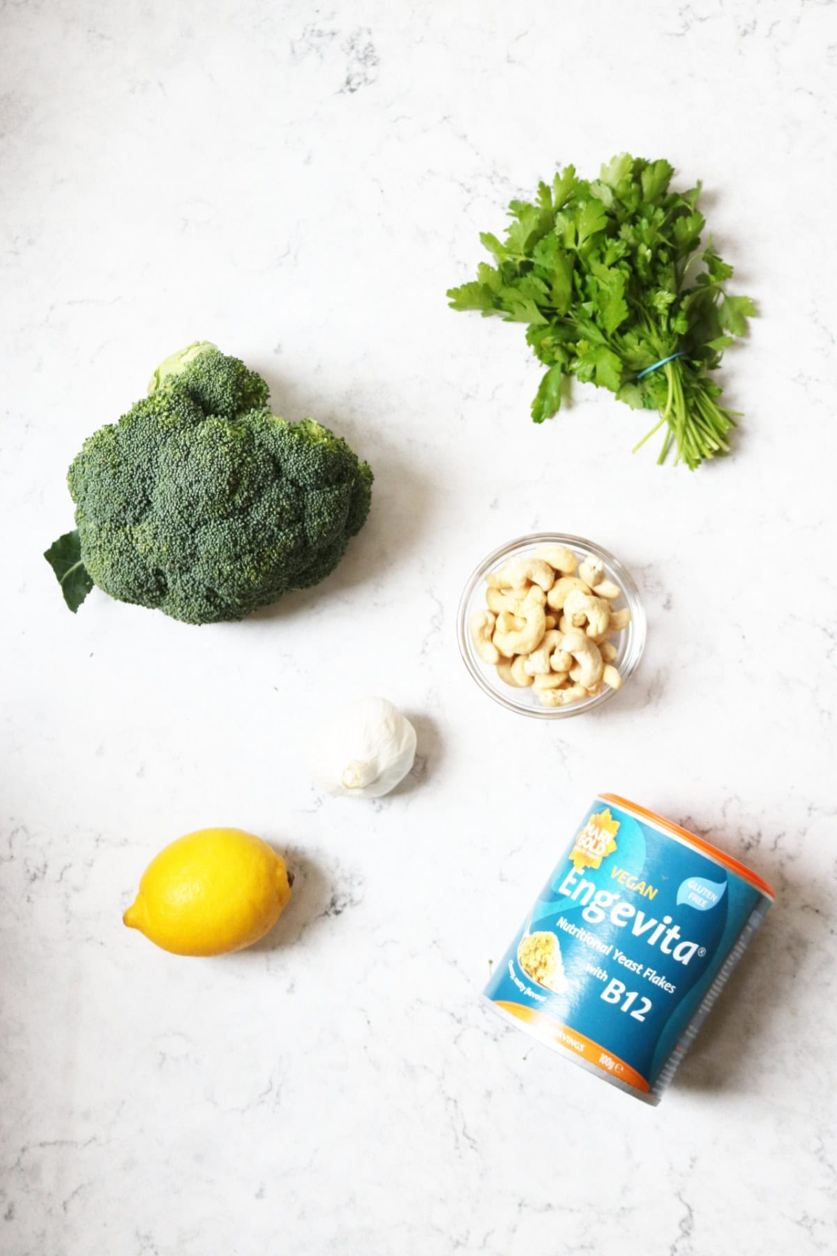 Parsley, broccoli, cashew nuts, garlic, lemon and nutritional yeast laid out on a white marble counter top