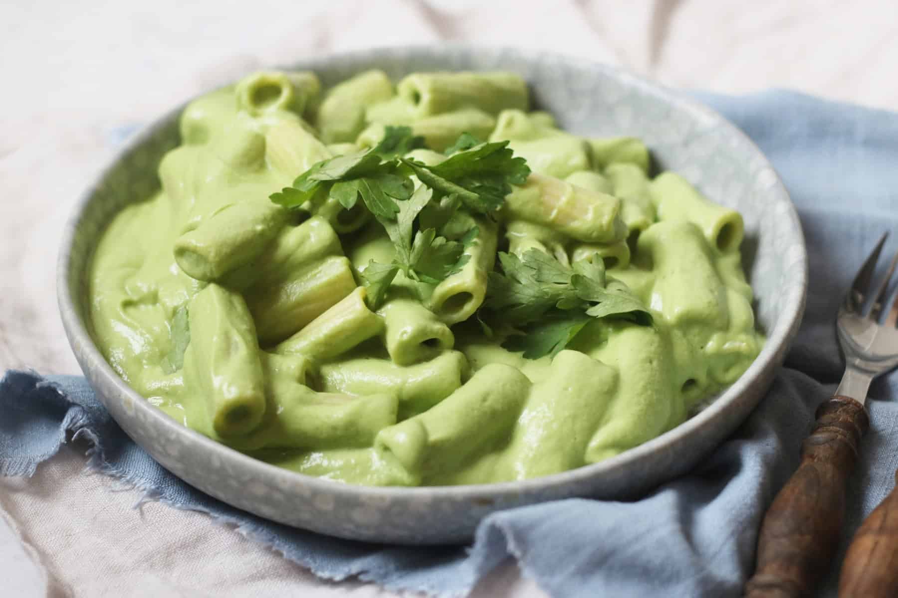 A plate of pasta made with Vegan Creamy Broccoli Pasta Sauce and garnished with parsley sat on top of a blue napkin with a knife and fork