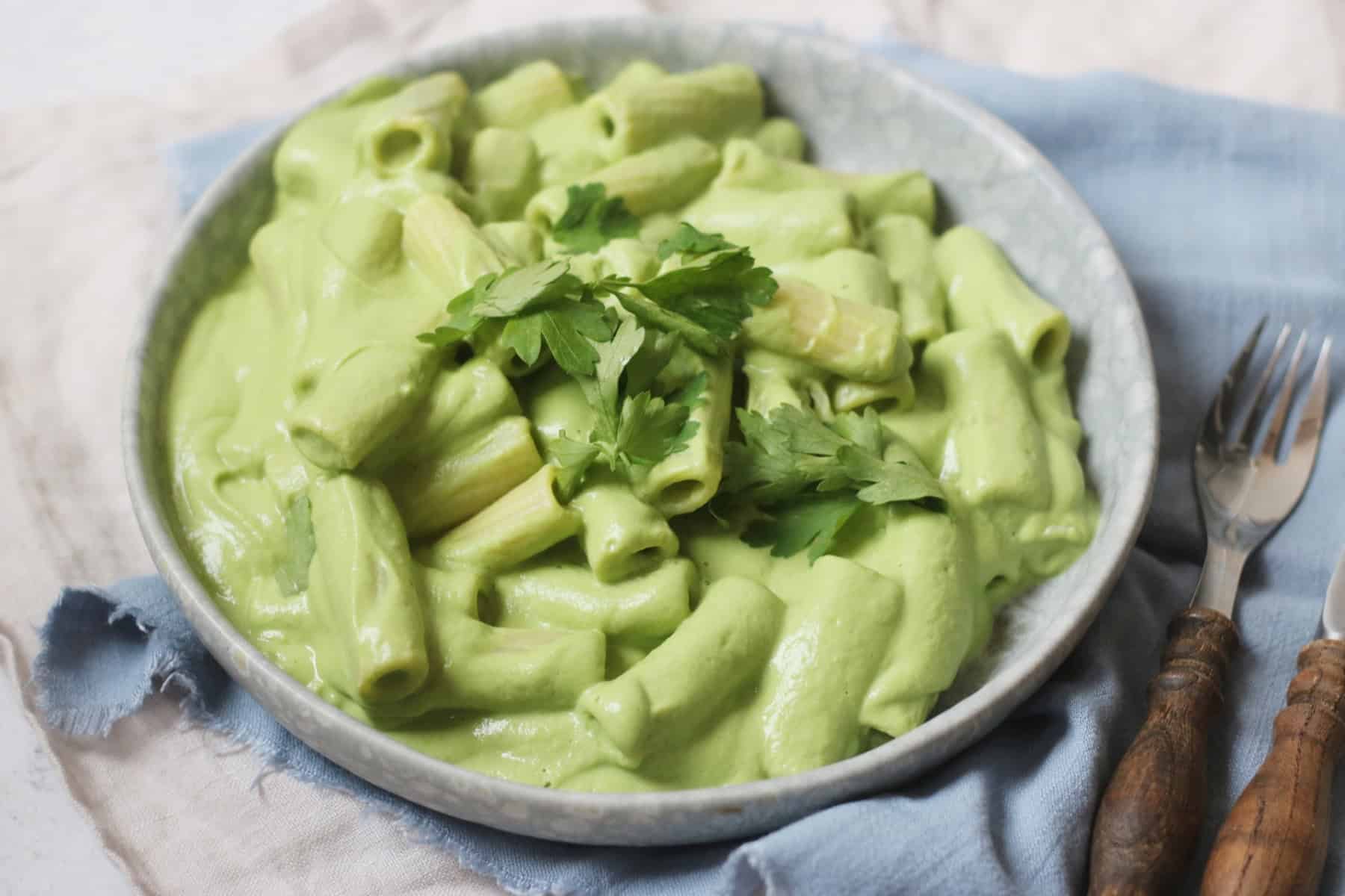 A plate of pasta made with Vegan Creamy Broccoli Pasta Sauce and garnished with parsley sat on top of a blue napkin with a knife and fork