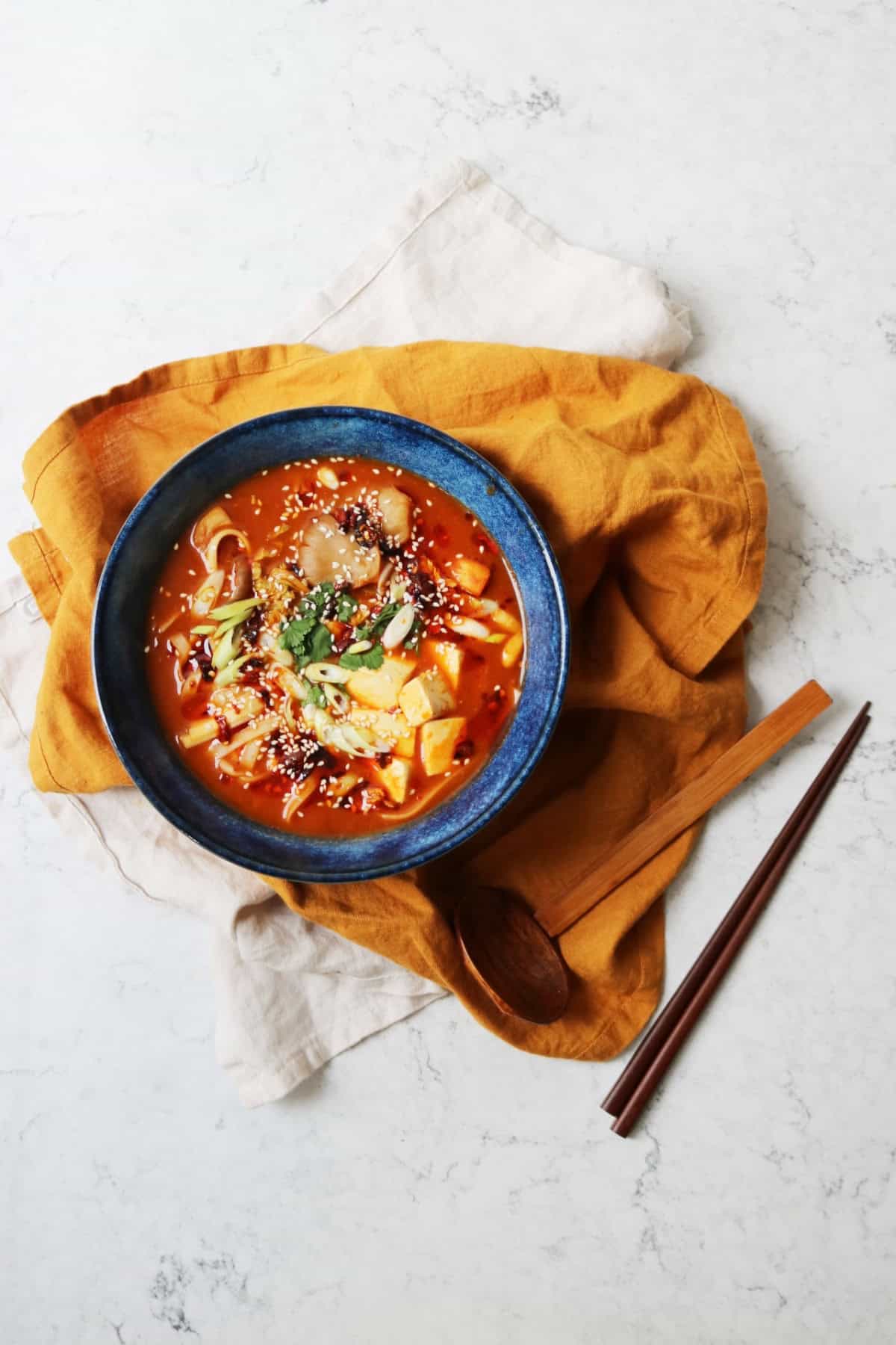 A bowl of Vegan Spicy Miso Ramen served in a blue bowl on top of a yellow napkin served alongside chopsticks and a spoon