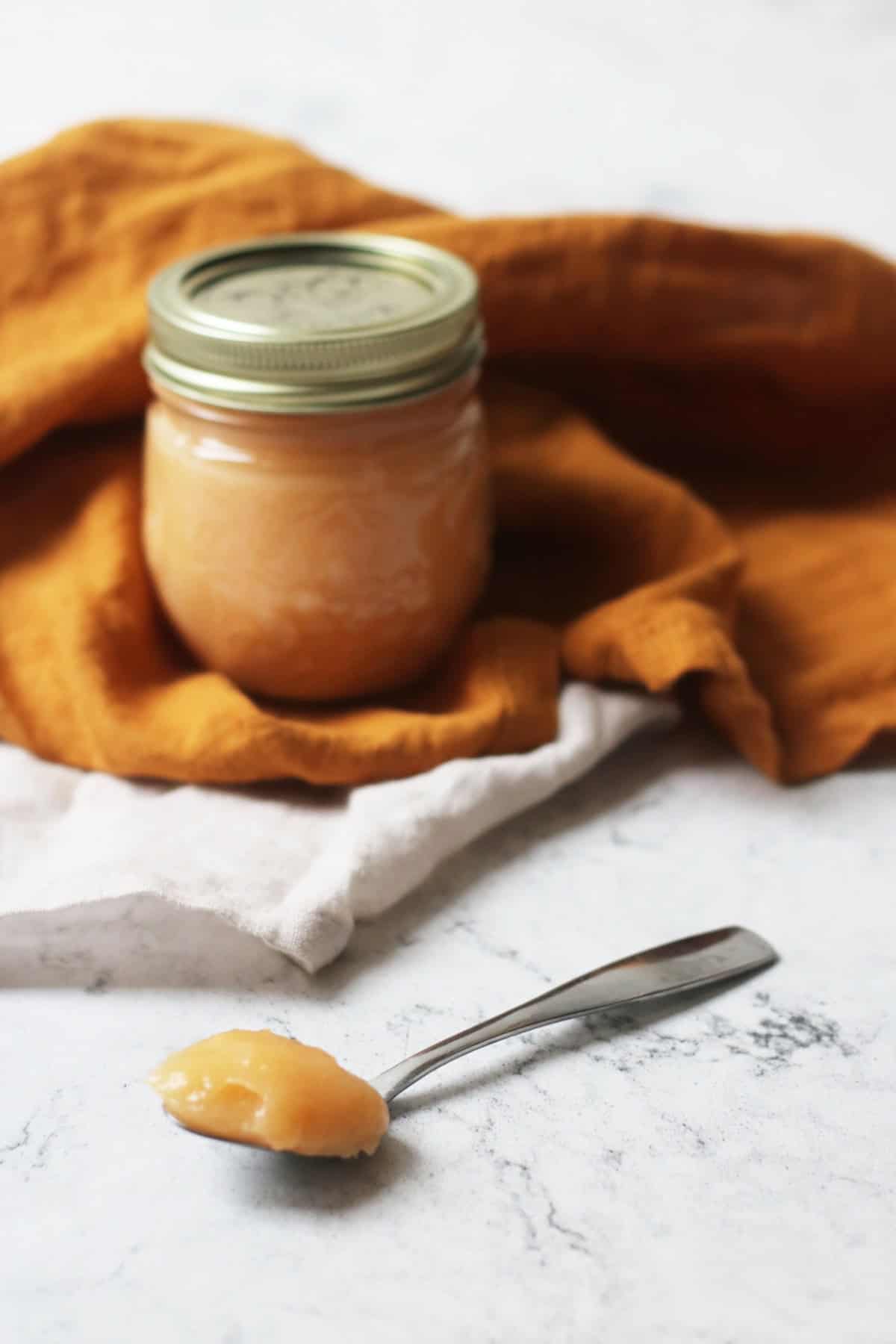 A jar of Vegan Blood Orange Curd nestled in an orange napkin with a spoon of the Vegan Blood Orange Curd resting on a white marble counter top