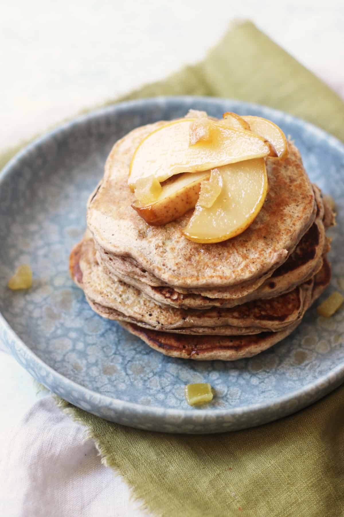 A close up of a stack of vegan pear and ginger pancakes topped with slices of caramelised pear and cubes of crystalised ginger on a blue plate with a green napkin and wooden handled knife and fork.