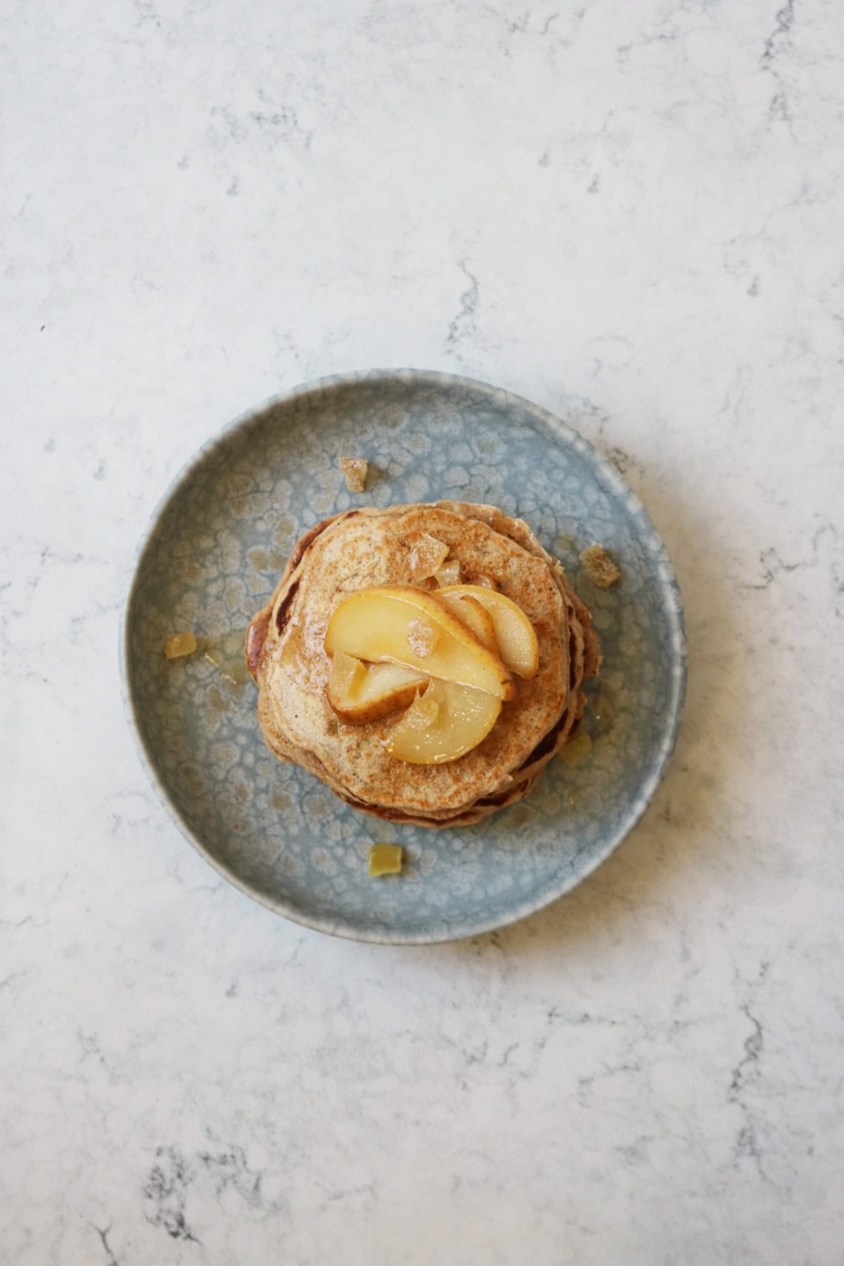 A stack of vegan pear and ginger pancakes topped with slices of caramelised pear and cubes of crystalised ginger on a blue plate seen from above.