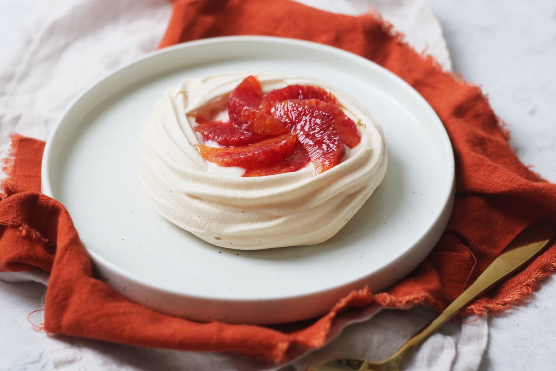 Vegan Blood Orange Pavlova on a white plate with orange napkin and gold spoon