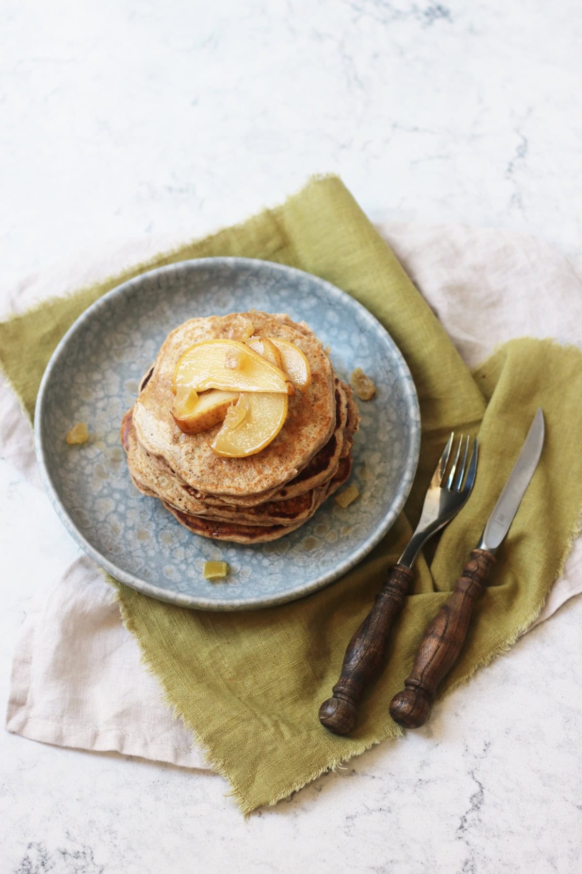 A stack of vegan pear and ginger pancakes topped with slices of caramelised pear and cubes of crystalised ginger on a blue plate with a green napkin and wooden handled knife and fork, seen from above.