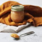 A jar of Vegan Blood Orange Curd nestled in an orange napkin with a spoon of the Vegan Blood Orange Curd resting on a white marble counter top