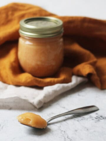 A jar of Vegan Blood Orange Curd nestled in an orange napkin with a spoon of the Vegan Blood Orange Curd resting on a white marble counter top