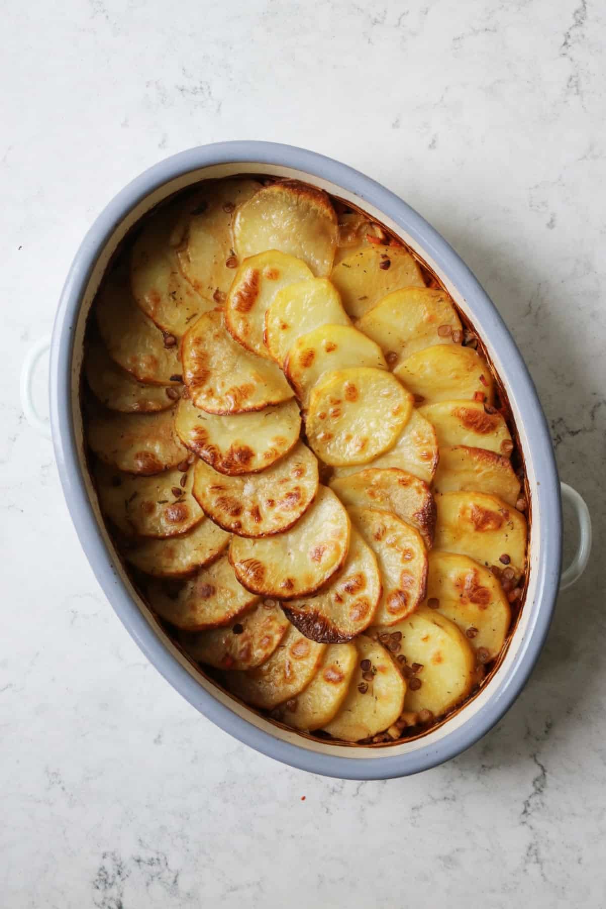 A Vegan Lancashire Hot Pot with crispy potato slices on top in an enamel oven dish placed on white marble counter top