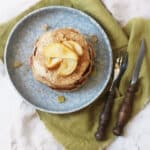 A stack of vegan pear and ginger pancakes topped with slices of caramelised pear and cubes of crystalised ginger seen from above, on a blue plate with a green napkin and wooden handled knife and fork.
