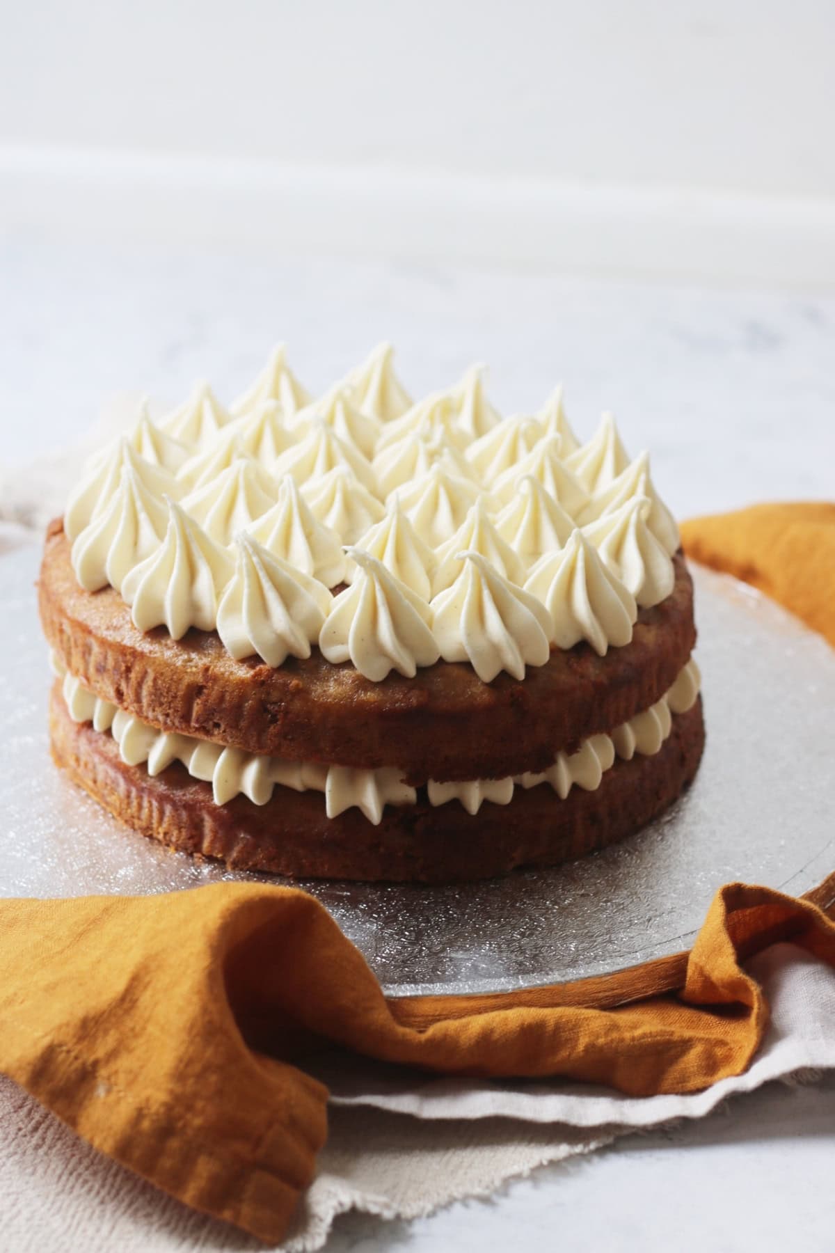 A Vegan Banana Cake with Vegan Cream Cheese Frosting on a silver cake board sat on top of yellow and cream napkins