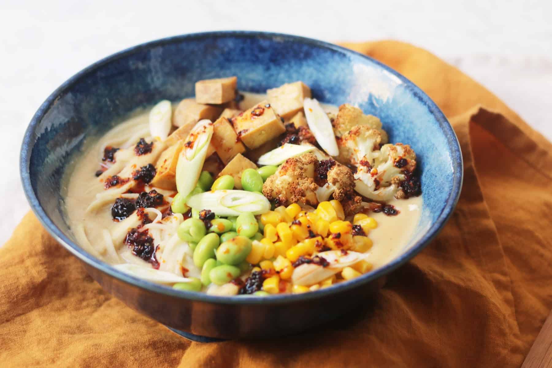 A bowl of Vegan Cauliflower Ramen topped with edamame, sweetcorn, roasted cauliflower, tofu, spring onions and chilli oil