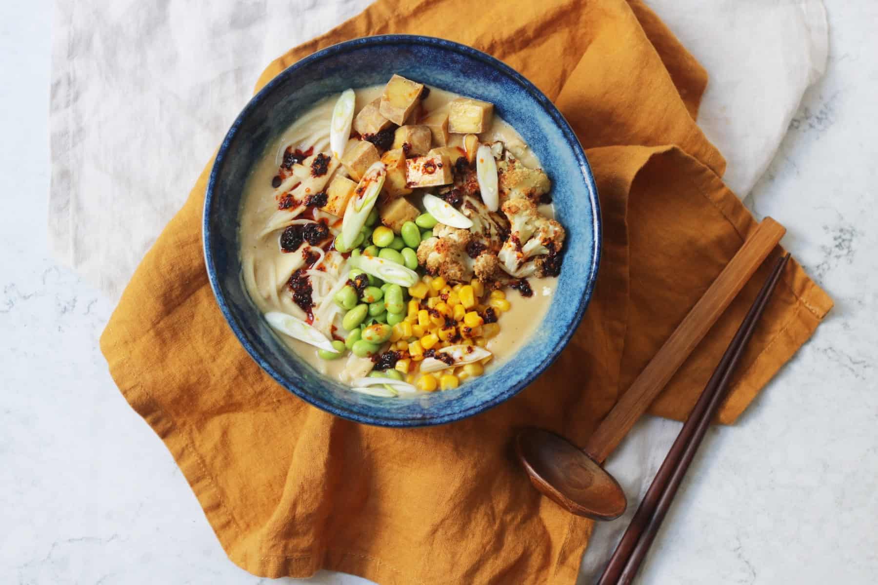 A bowl of Vegan Cauliflower Ramen topped with edamame, sweetcorn, roasted cauliflower, tofu, spring onions and chilli oil served with wooden chopsticks on top of a yellow napkin