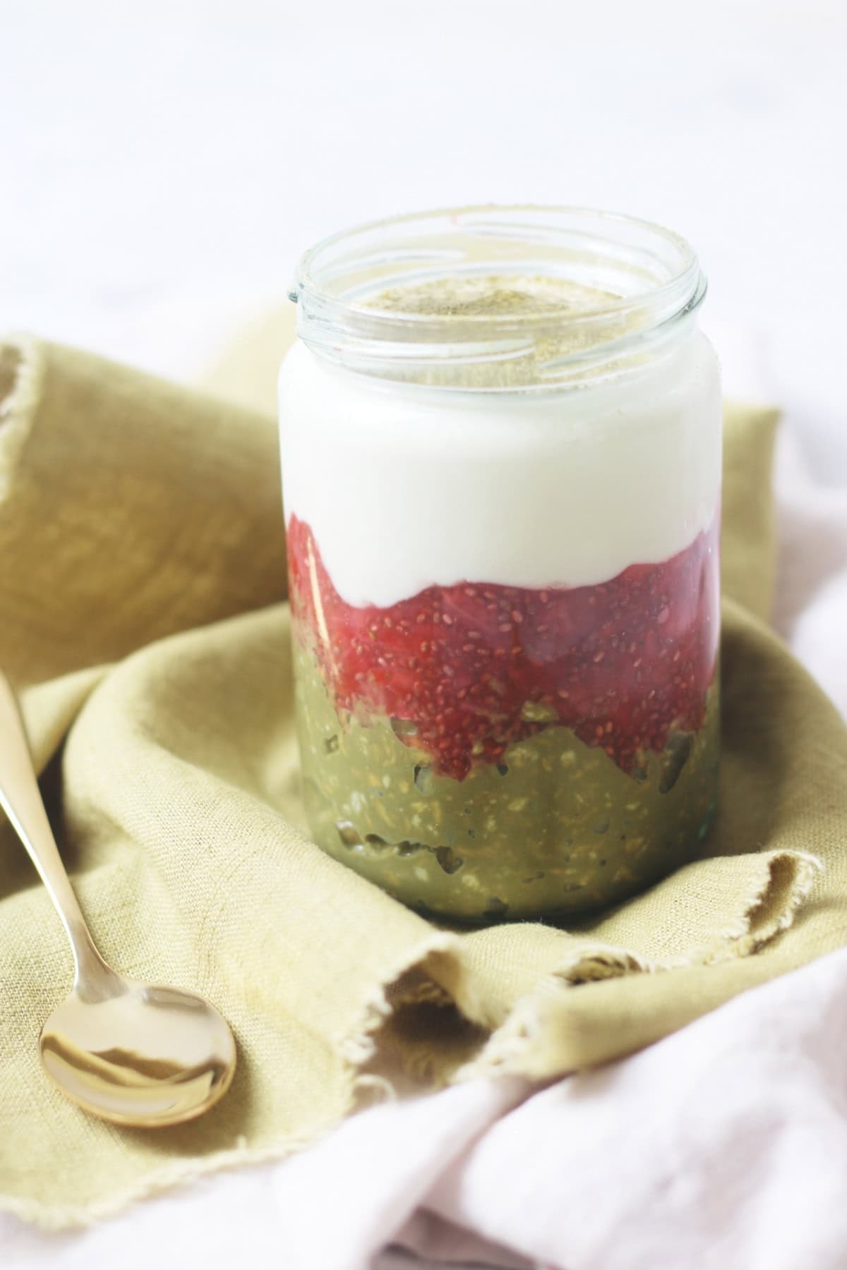 A jar of Vegan Strawberry and Matcha Overnight Oats on a green napkin with a gold teaspoon