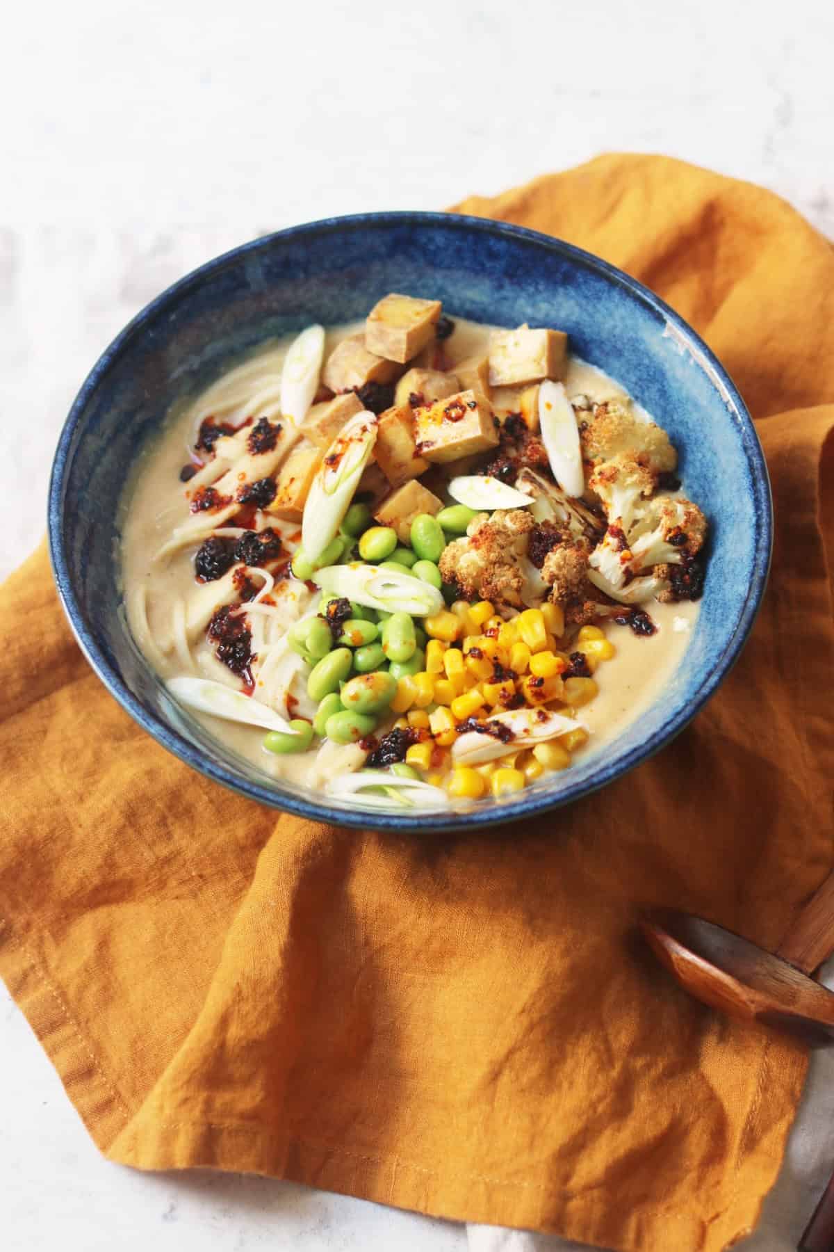 A bowl of Vegan Cauliflower Ramen topped with edamame, sweetcorn, roasted cauliflower, tofu, spring onions and chilli oil served with wooden chopsticks on top of a yellow napkin