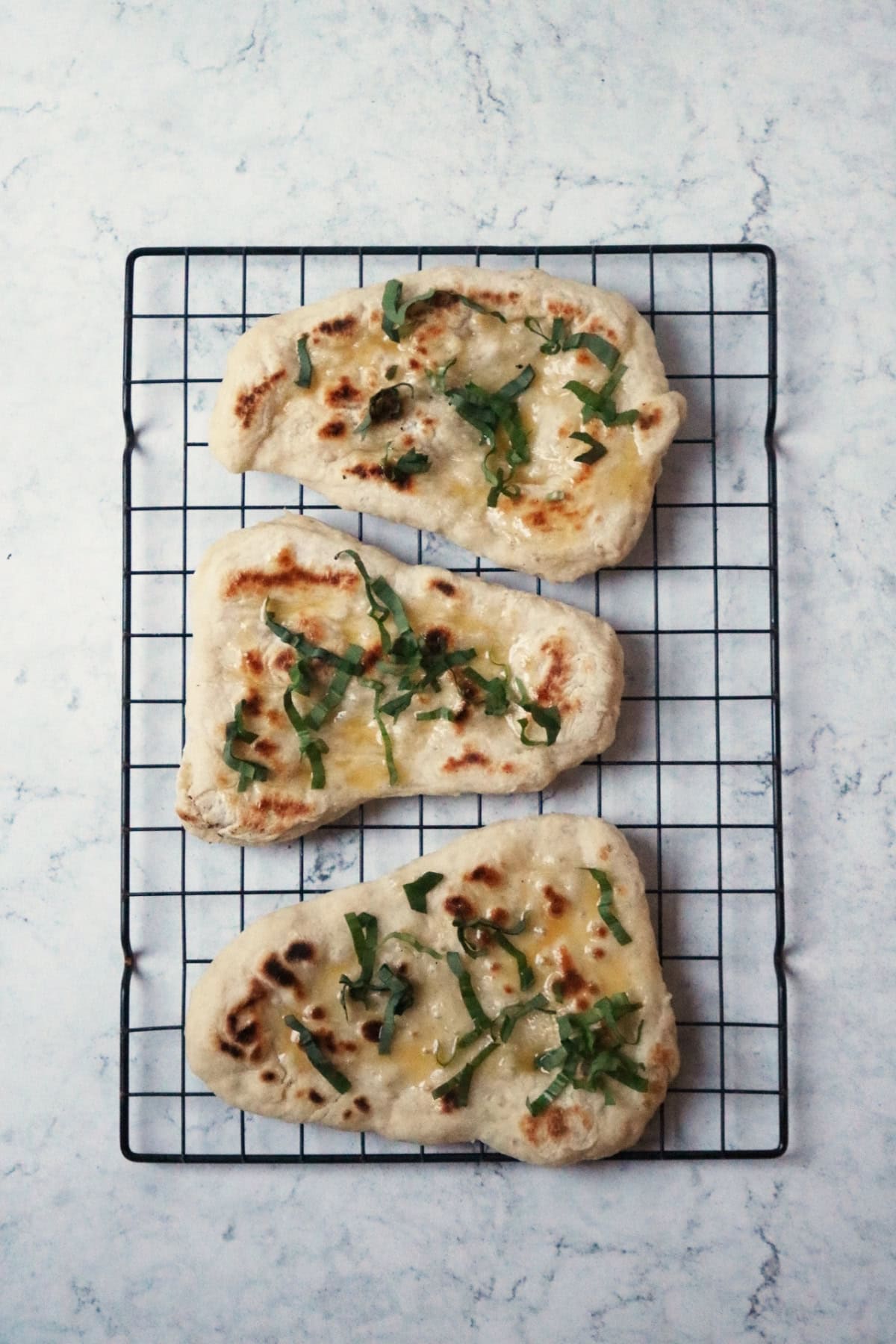 3 Vegan Wild Garlic Naan Breads cooling on a wire rack. The naan breads have been liberally brushed with melted vegan butter and topped with ribbons of wild garlic