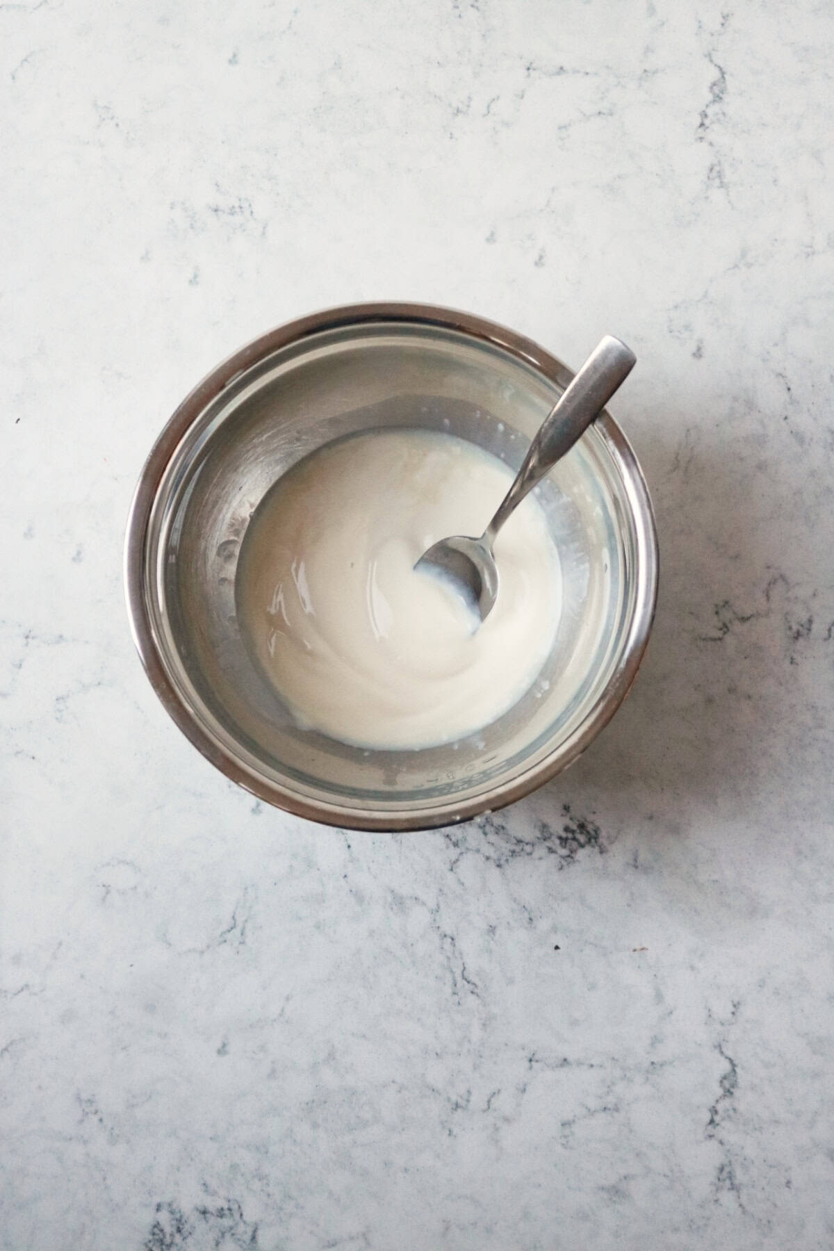 A small metal mixing bowl with a yogurt based dressing being stirred by a metal spoon