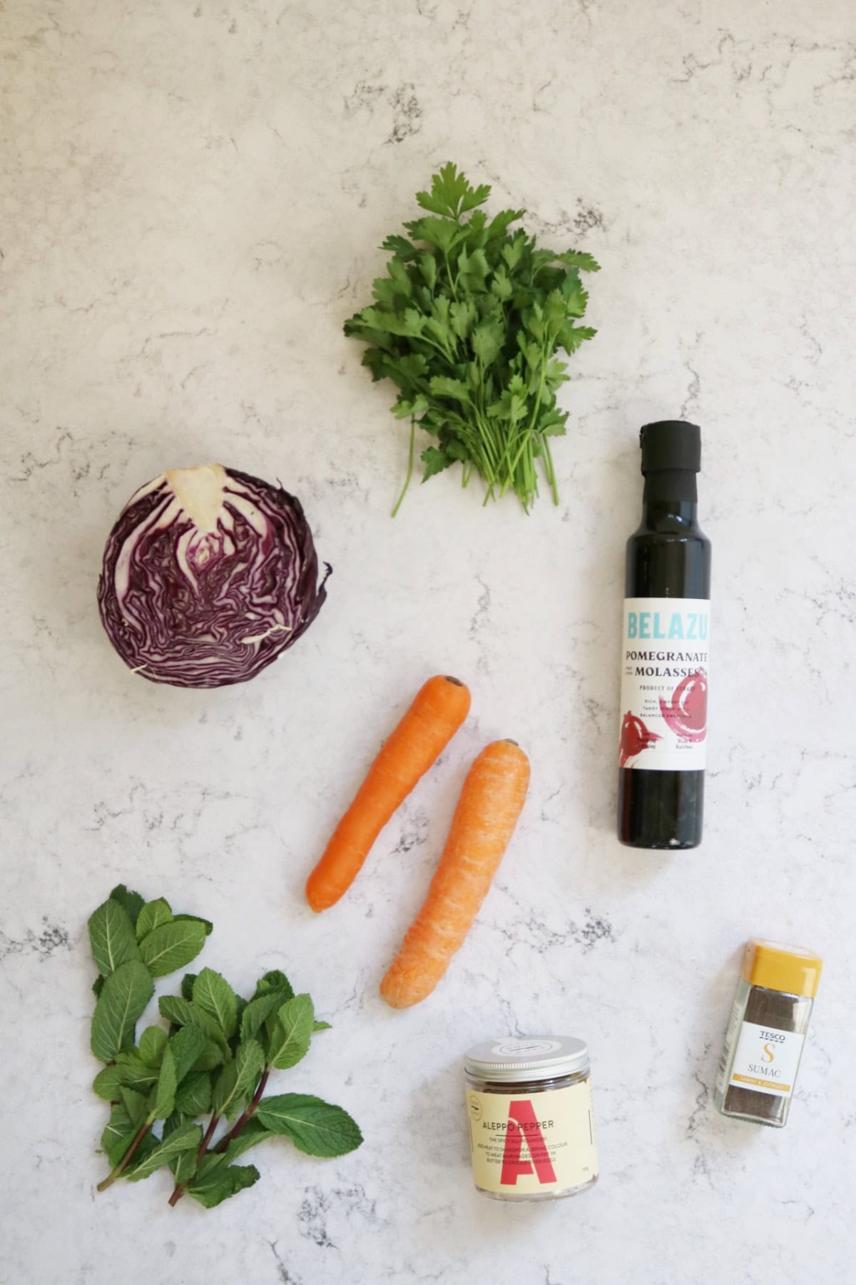 A bunch of parsley, half a red cabbage, a bottle of pomegranate molasses, two carrots, a bunch of mint, Aleppo pepper and sumac laid out on a white marble counter top
