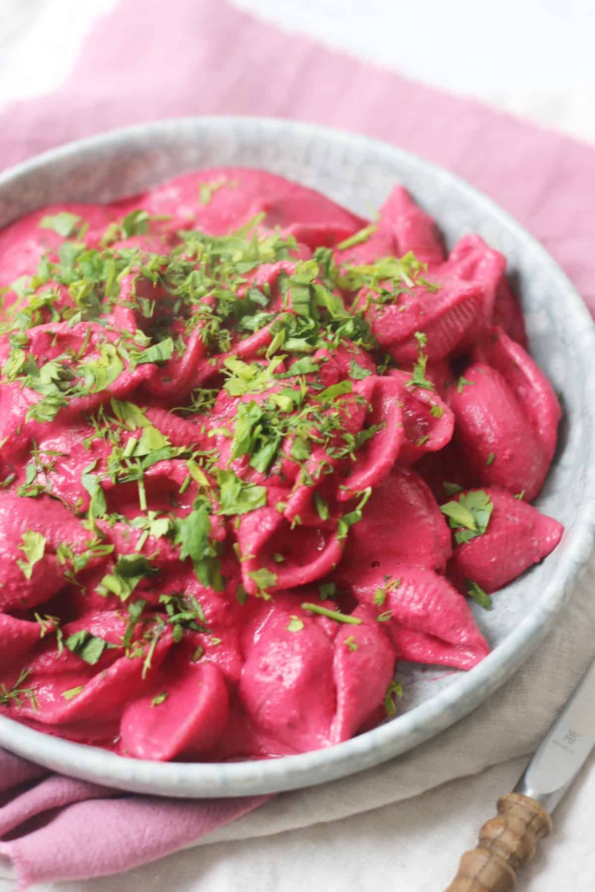 Vegan Creamy Beetroot Pasta Sauce served with pasta and fresh herbs on a pale blue plate with wooden handled knife and fork.