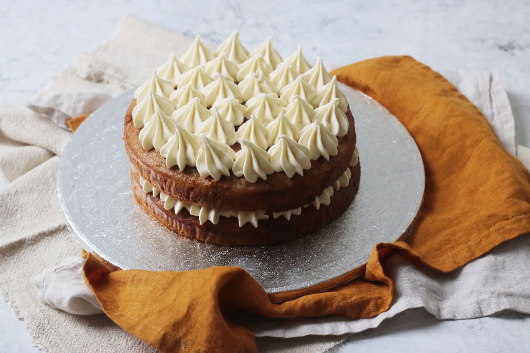 A Vegan Banana Cake with Vegan Cream Cheese Frosting on a silver cake board sat on top of yellow and cream napkins