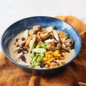 A bowl of Vegan Cauliflower Ramen topped with edamame, sweetcorn, roasted cauliflower, tofu, spring onions and chilli oil on top of a yellow napkin