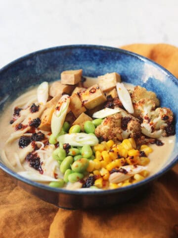 A bowl of Vegan Cauliflower Ramen topped with edamame, sweetcorn, roasted cauliflower, tofu, spring onions and chilli oil on top of a yellow napkin