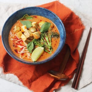 Vegan Thai Red Curry Noodle Soup served in a dark blue ramen bowl with an orange napkin and wood spoons and chopsticks