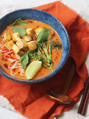 Vegan Thai Red Curry Noodle Soup served in a dark blue ramen bowl with an orange napkin and wood spoons and chopsticks