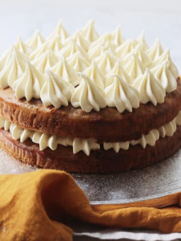 A Vegan Banana Cake with Vegan Cream Cheese Frosting on a silver cake board sat on top of yellow and cream napkins