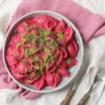 Vegan Creamy Beetroot Pasta Sauce served with pasta and fresh herbs on a pale blue plate with wooden handled knife and fork.