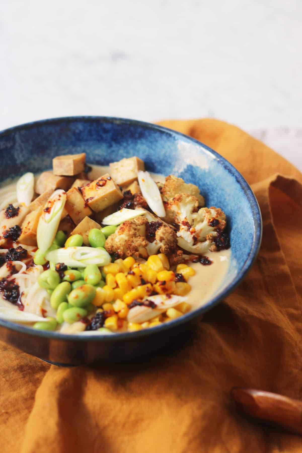 A bowl of Vegan Cauliflower Ramen topped with edamame, sweetcorn, roasted cauliflower, tofu, spring onions and chilli oil on top of a yellow napkin