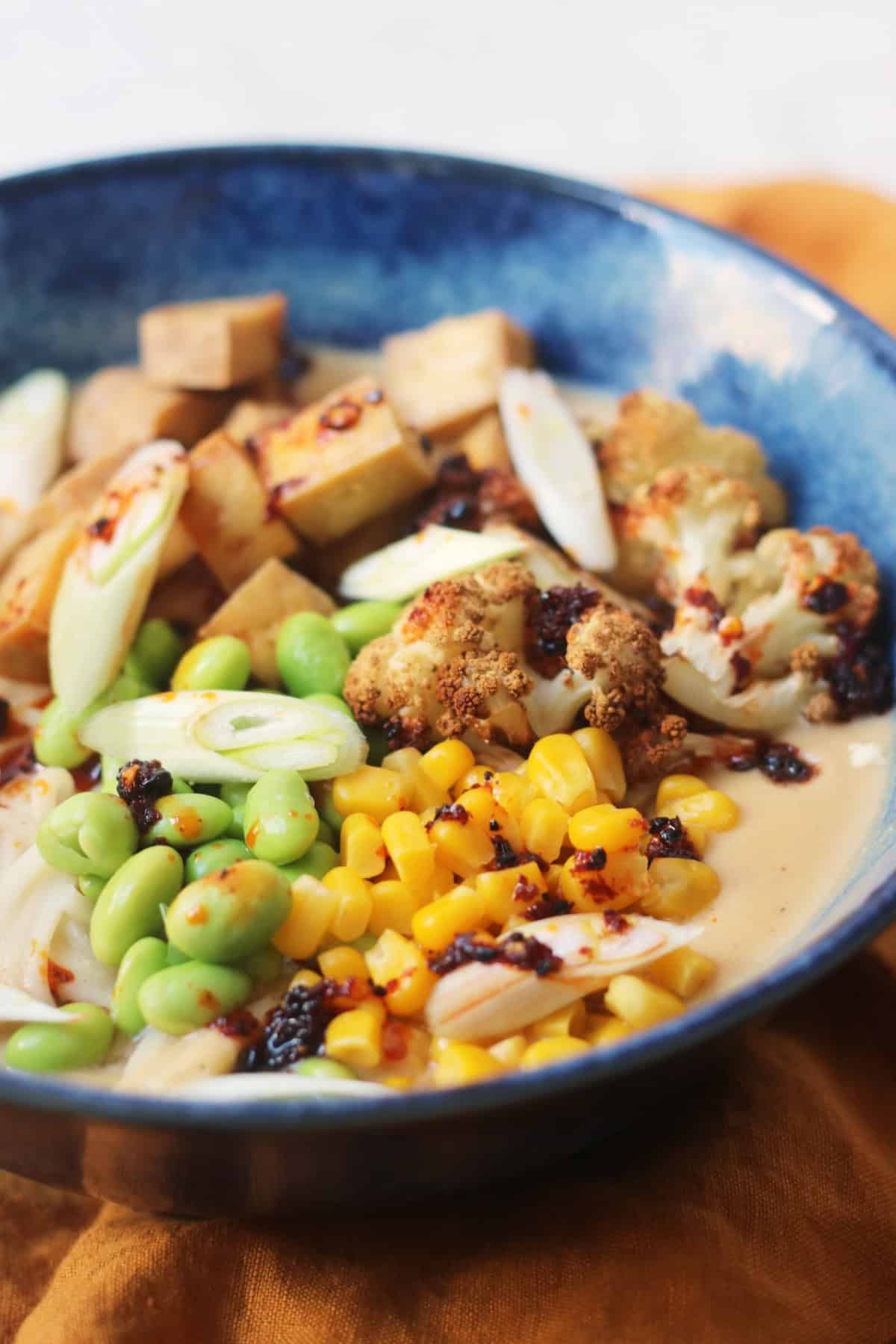 A bowl of Vegan Cauliflower Ramen topped with edamame, sweetcorn, roasted cauliflower, tofu, spring onions and chilli oil