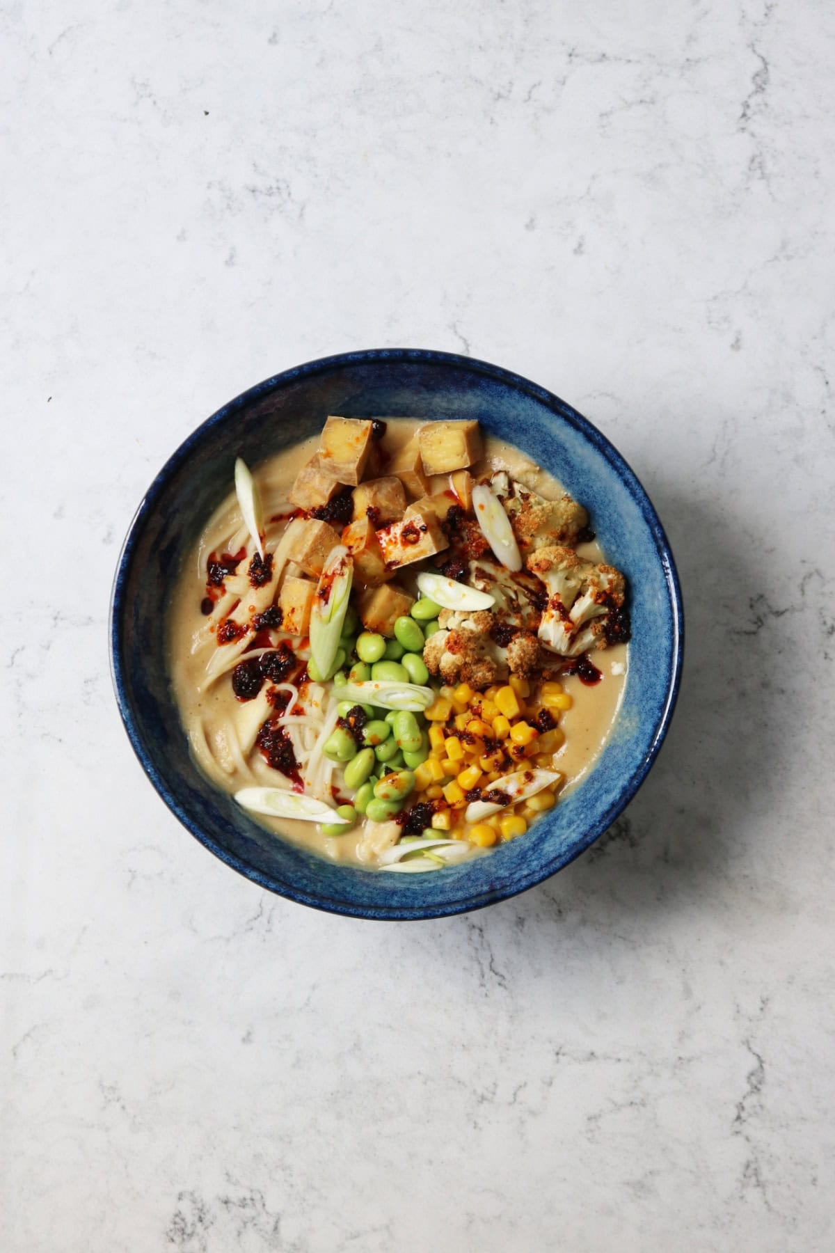 A bowl of Vegan Cauliflower Ramen topped with edamame, sweetcorn, roasted cauliflower, tofu, spring onions and chilli oil