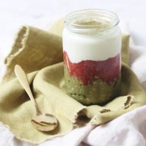 A jar of Vegan Strawberry and Matcha Overnight Oats on a green napkin with a gold teaspoon