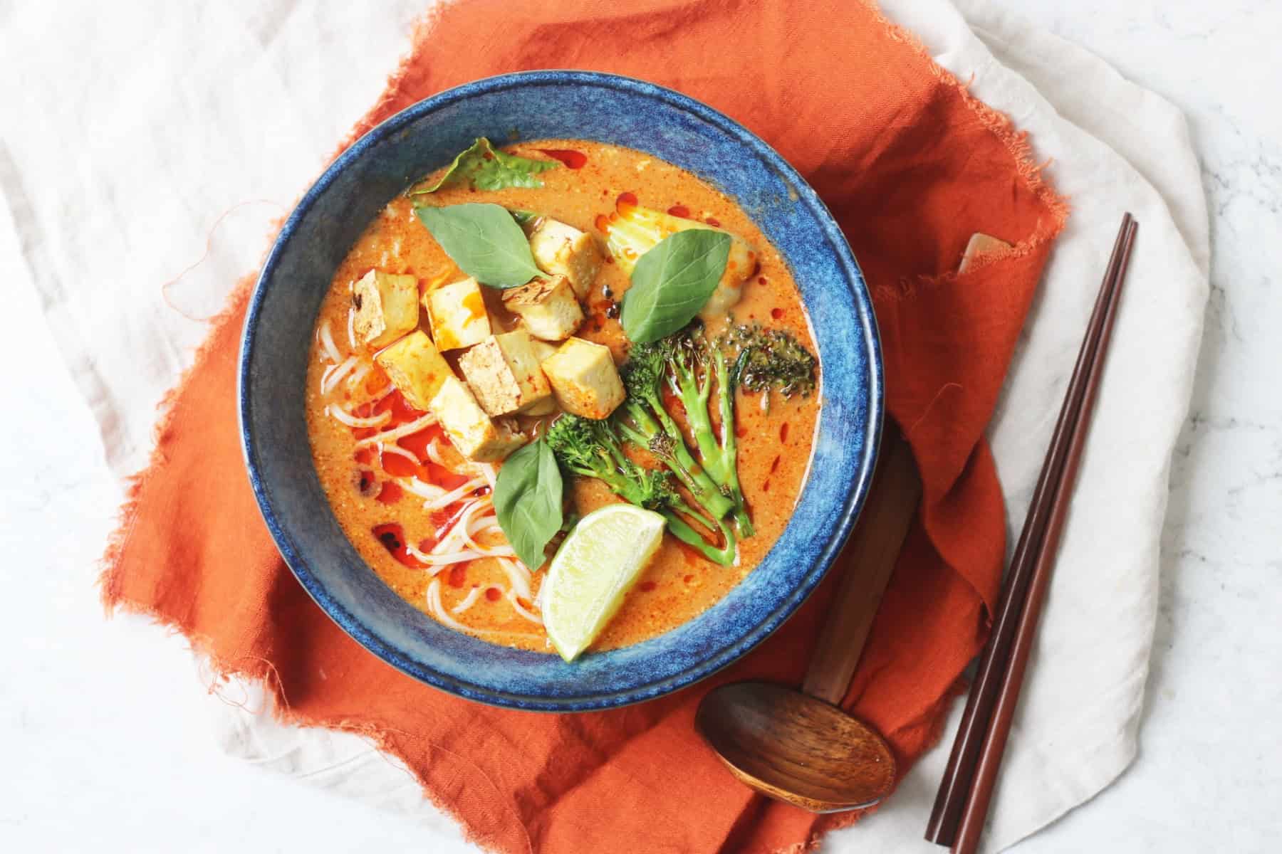 Vegan Thai Red Curry Noodle Soup served in a dark blue ramen bowl with an orange napkin and wood spoons and chopsticks