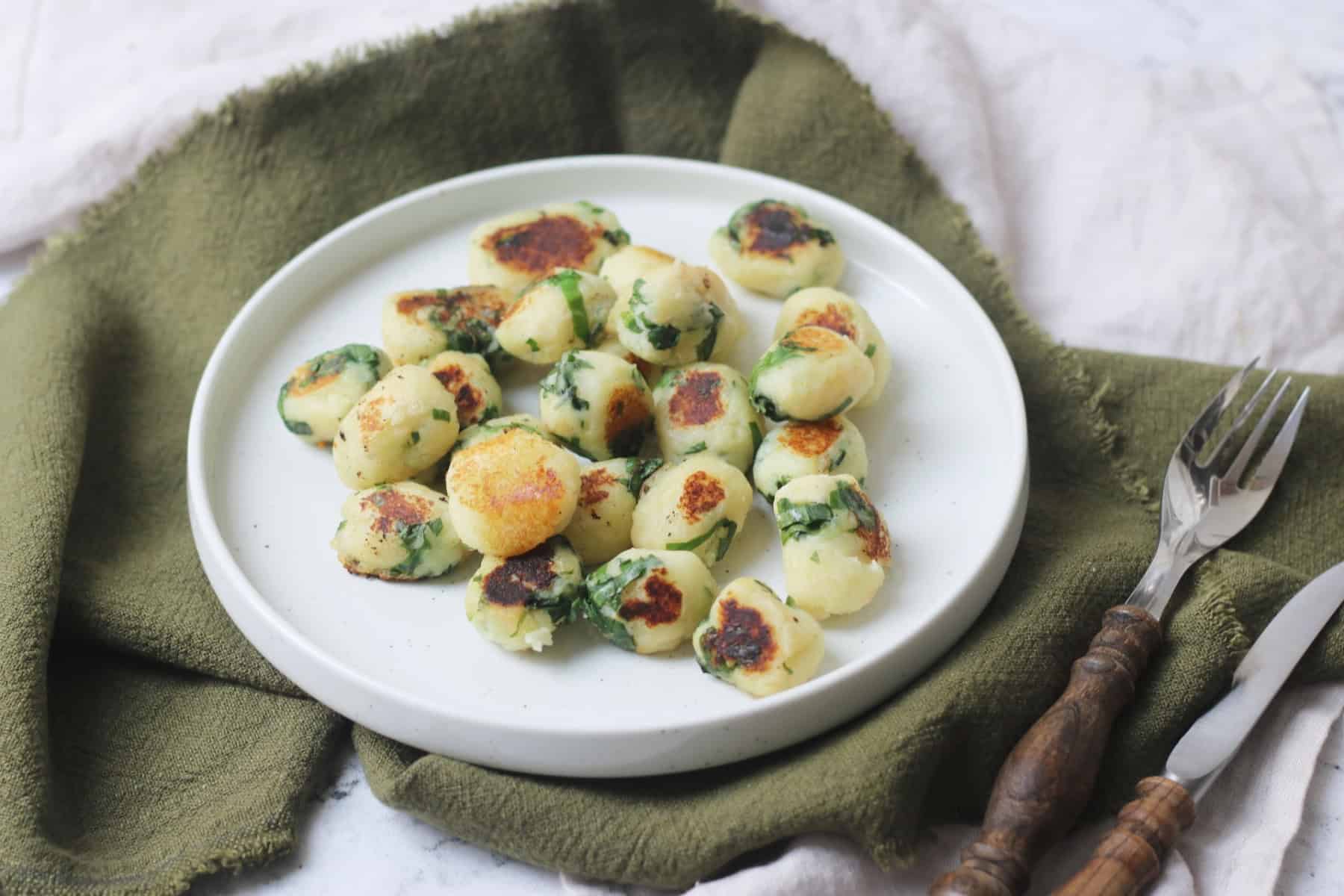 A portion of Vegan Wild Garlic Gnocchi on a white plate that is sat on top of a dark green and cream napkin with a wooden handled knife and fork