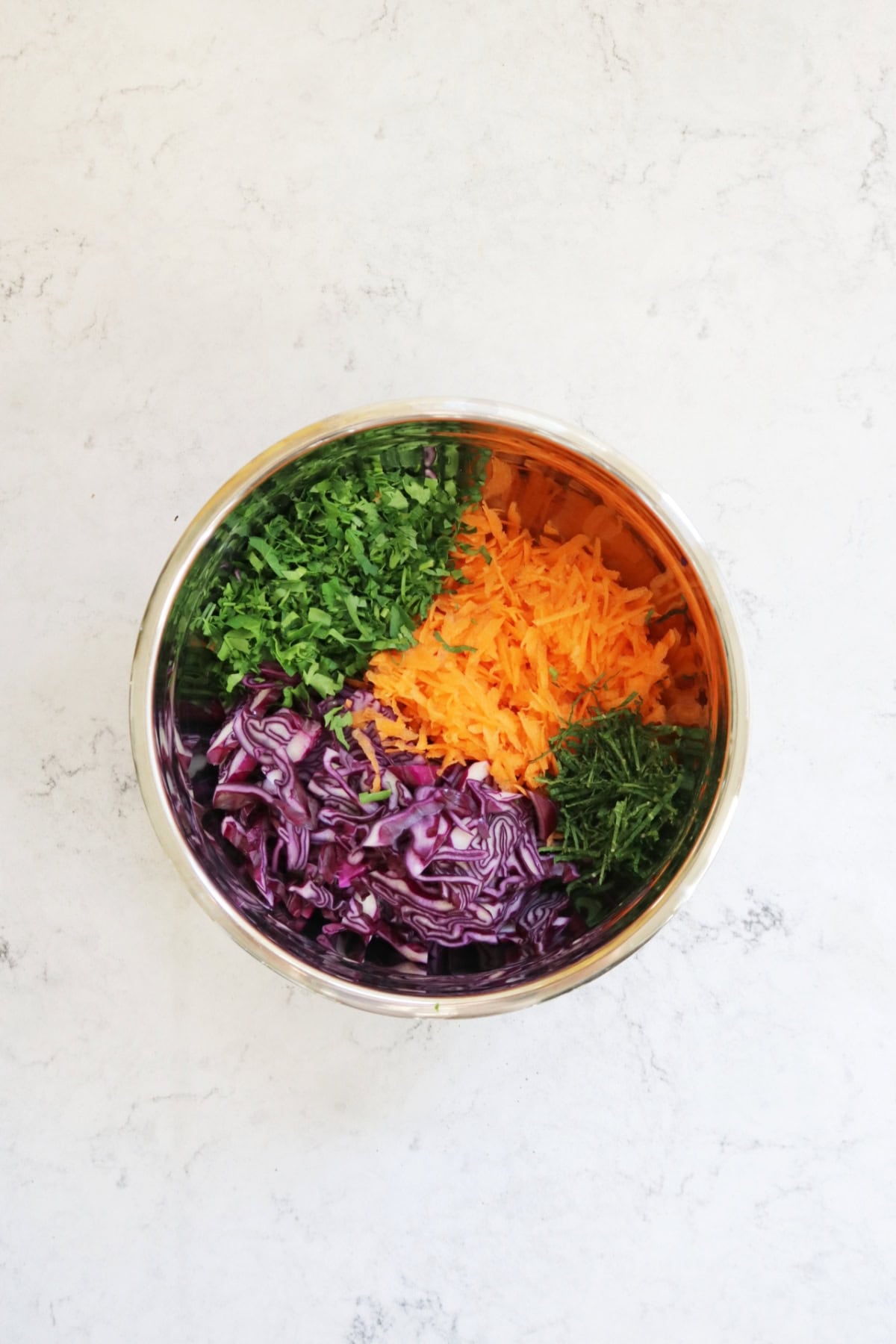 A metal mixing bowl containing shredded red cabbage, grated carrot and chopped parsley and mint