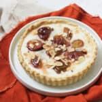 A vegan plum and custard tart topped with toasted flaked almonds on a white plate with a dark orange napkin.