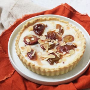 A vegan plum and custard tart topped with toasted flaked almonds on a white plate with a dark orange napkin.