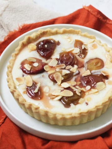 A vegan plum and custard tart topped with toasted flaked almonds on a white plate with a dark orange napkin.