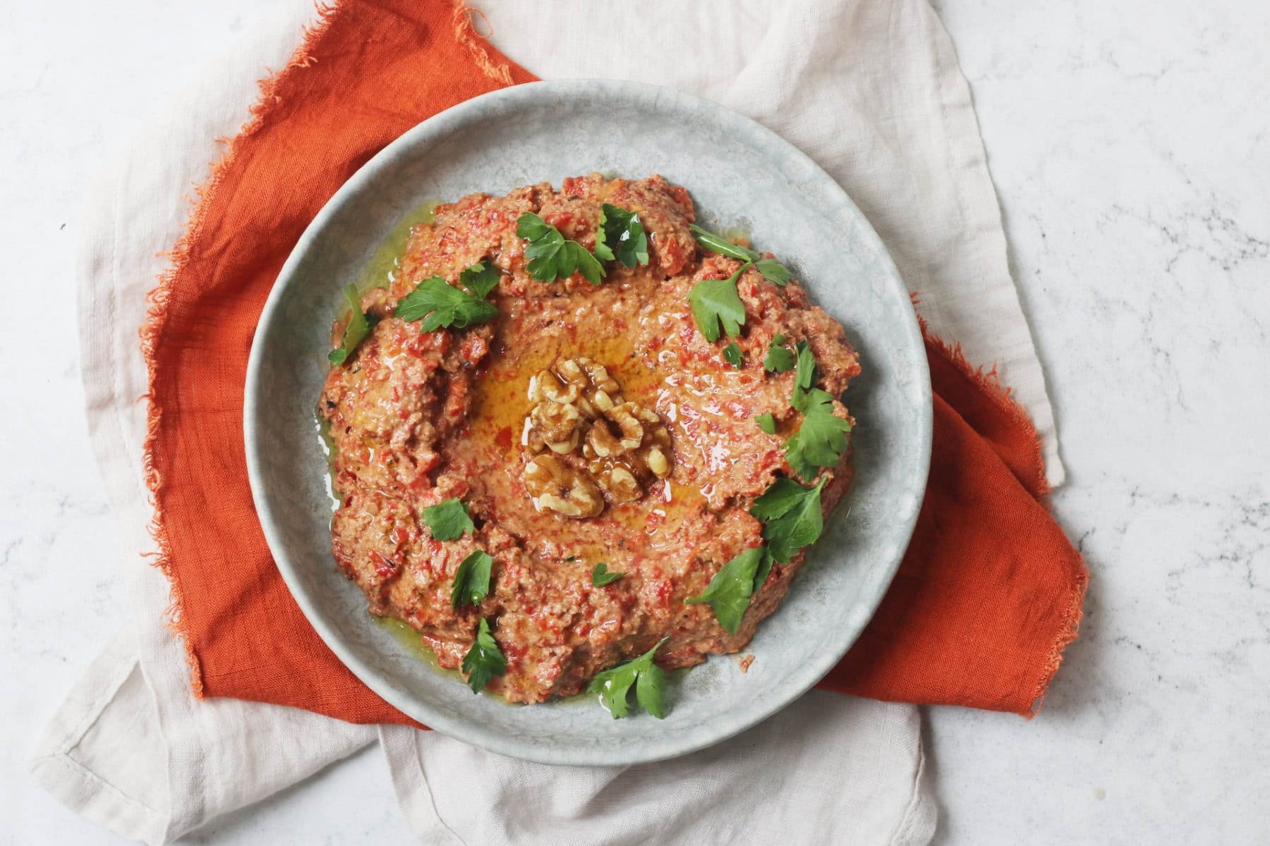Muhammara, a roasted red pepper and walnut dip served with walnut halves and parsley on a pale blue plate with an orange napkin