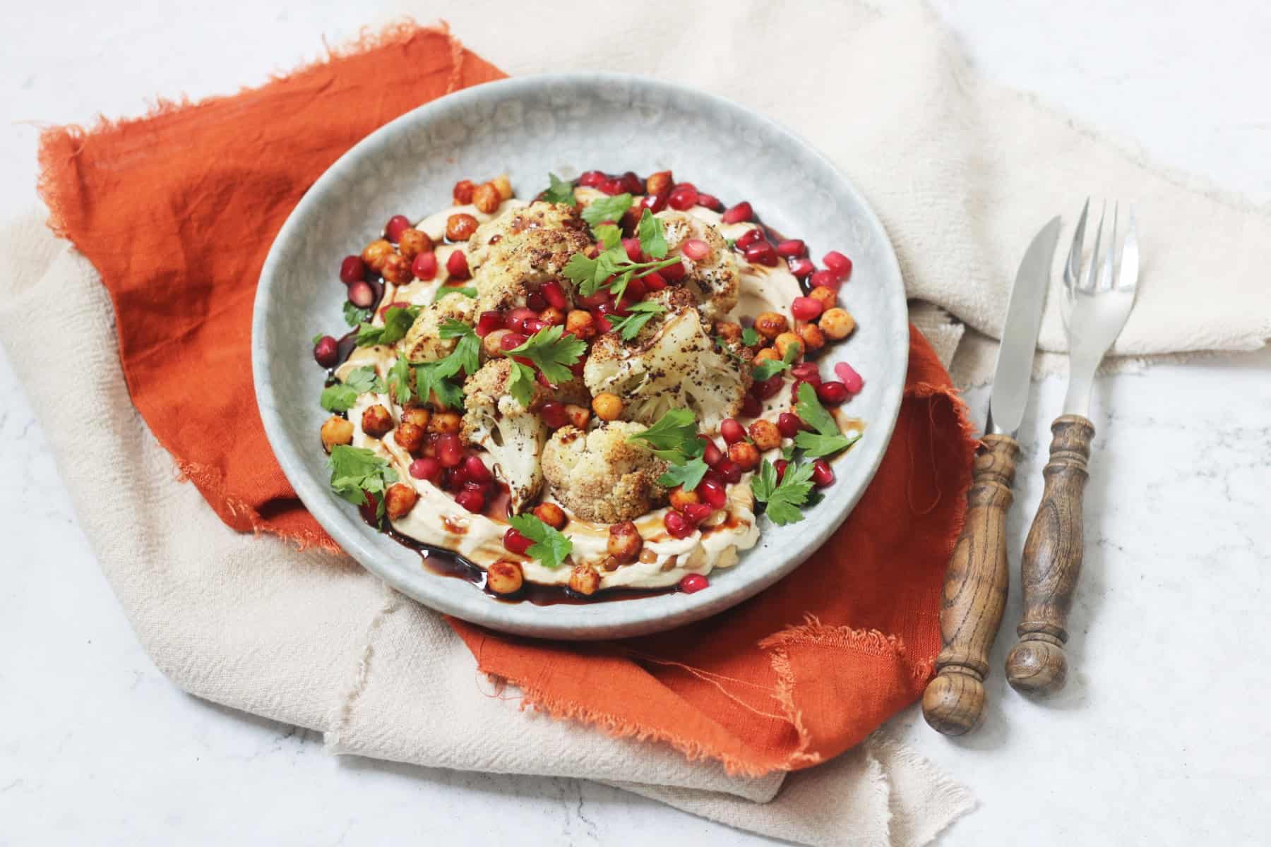 A plate of sumac roasted cauliflower on a bed of hummus with a drizzling of pomegranate molasses and roasted chickpeas, pomegranate seeds and parsley scattered over the top