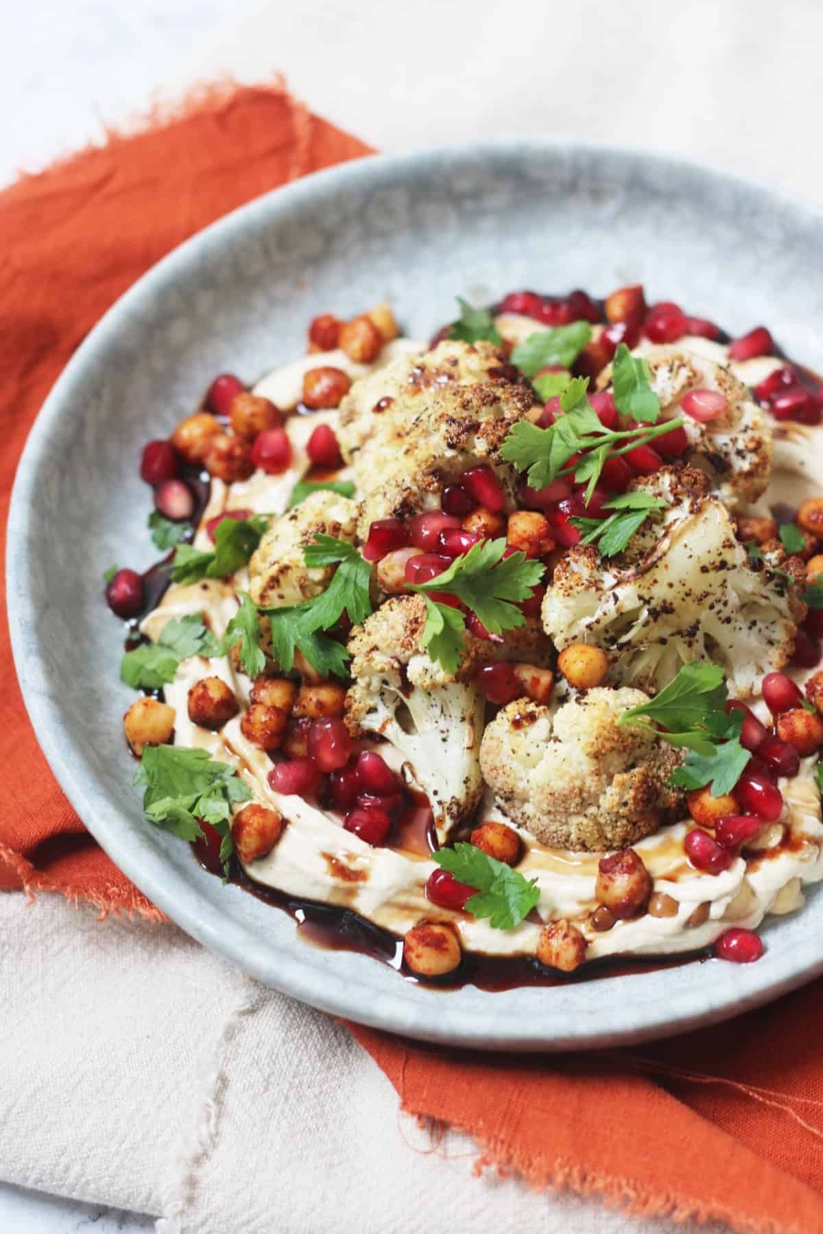 A plate of sumac roasted cauliflower on a bed of hummus with a drizzling of pomegranate molasses and roasted chickpeas, pomegranate seeds and parsley scattered over the top