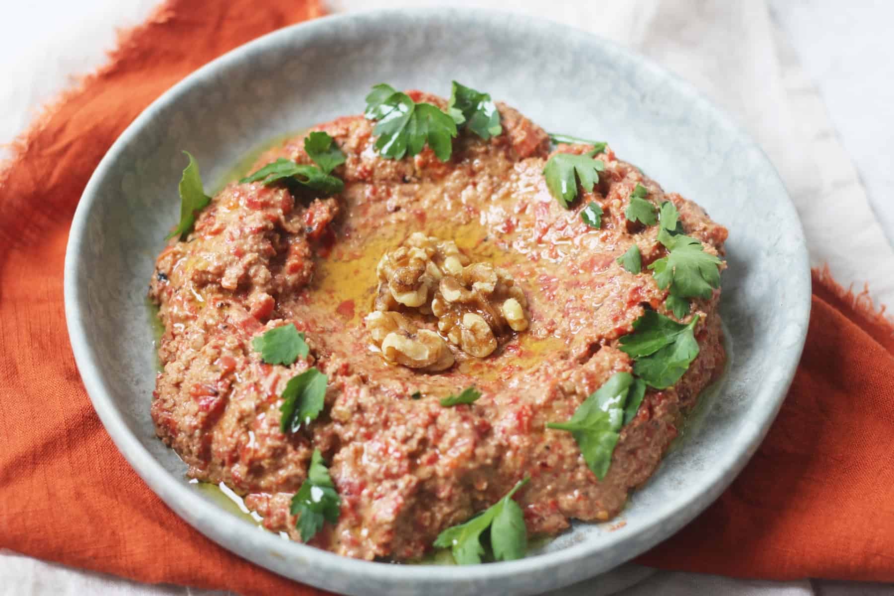Muhammara, a roasted red pepper and walnut dip served with walnut halves and parsley on a pale blue plate with an orange napkin