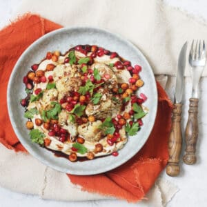 A plate of sumac roasted cauliflower on a bed of hummus with a drizzling of pomegranate molasses and roasted chickpeas, pomegranate seeds and parsley scattered over the top