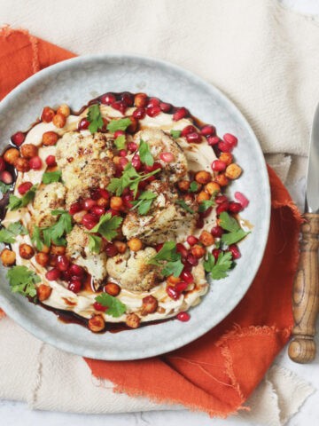 A plate of sumac roasted cauliflower on a bed of hummus with a drizzling of pomegranate molasses and roasted chickpeas, pomegranate seeds and parsley scattered over the top
