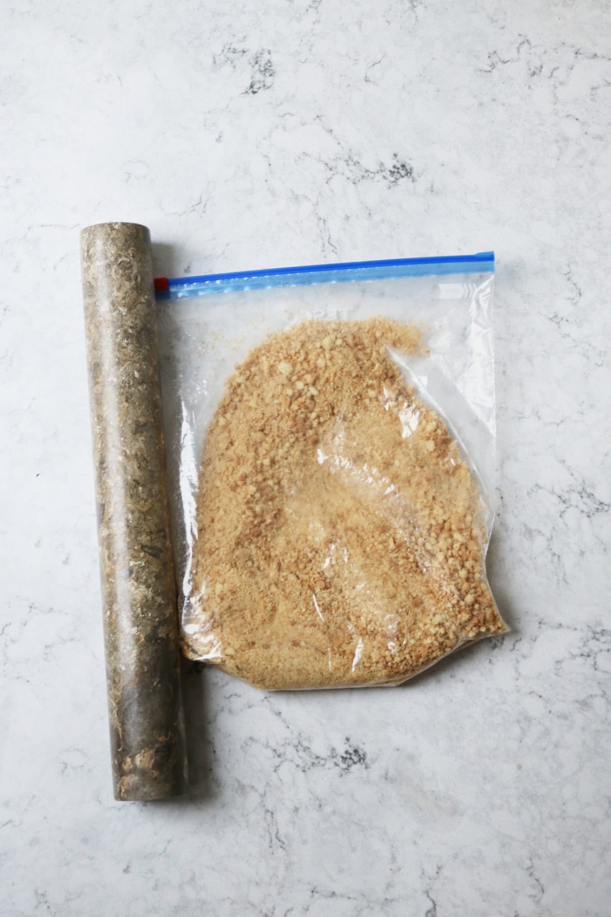 A Ziploc bag filled with crushed biscuit crumbs and a grey marble rolling pin sat on a white marble counter top