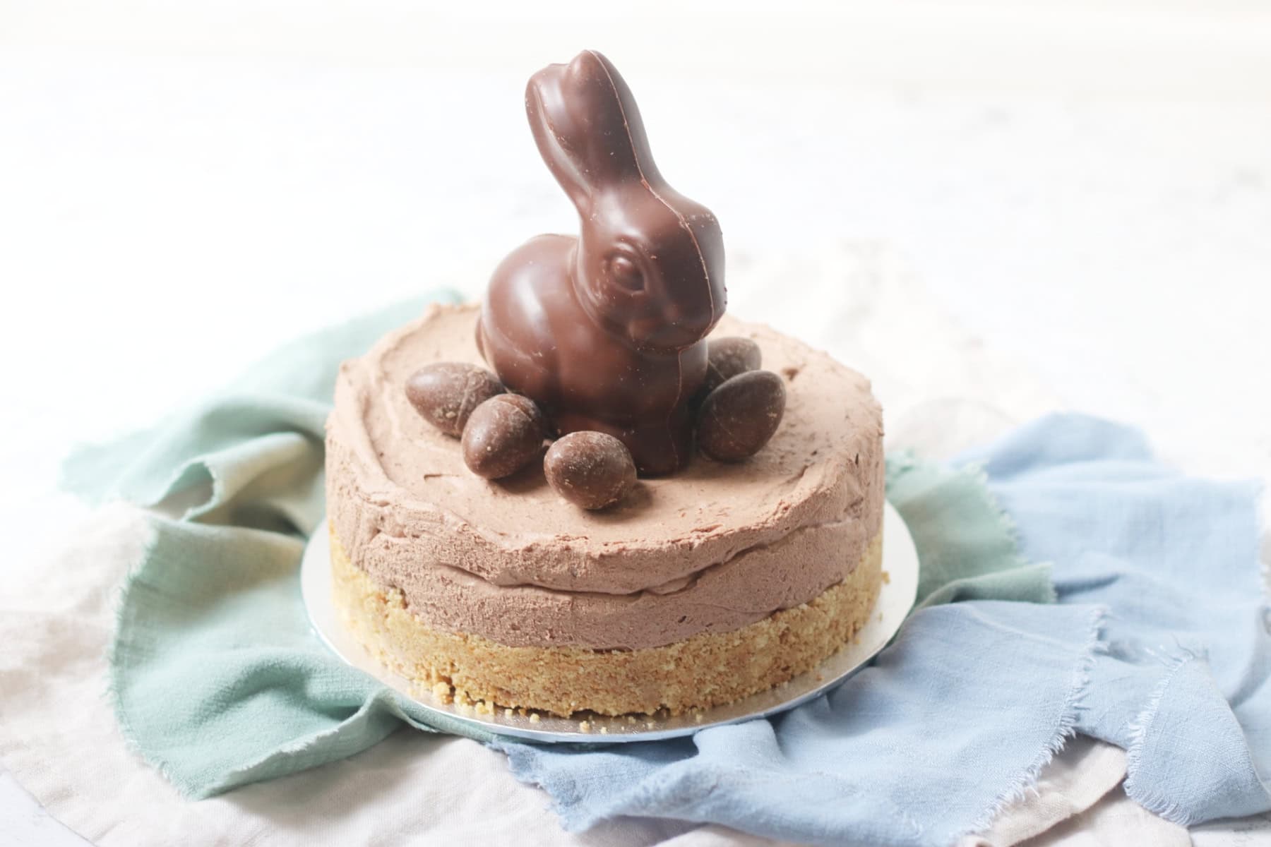 A Vegan Chocolate Cheesecake decorated with a chocolate bunny rabbit and small chocolate eggs sat on top of a silver cake board and blue and turquoise napkins