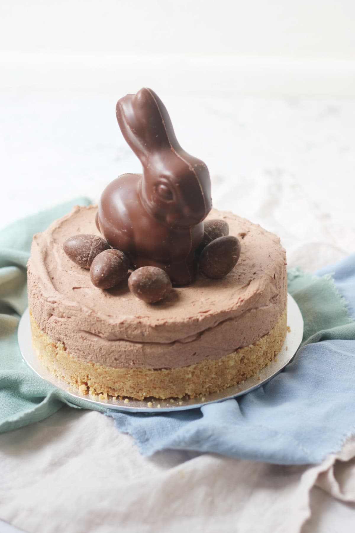 A Vegan Chocolate Cheesecake decorated with a chocolate bunny rabbit and small chocolate eggs sat on top of a silver cake board and blue and turquoise napkins