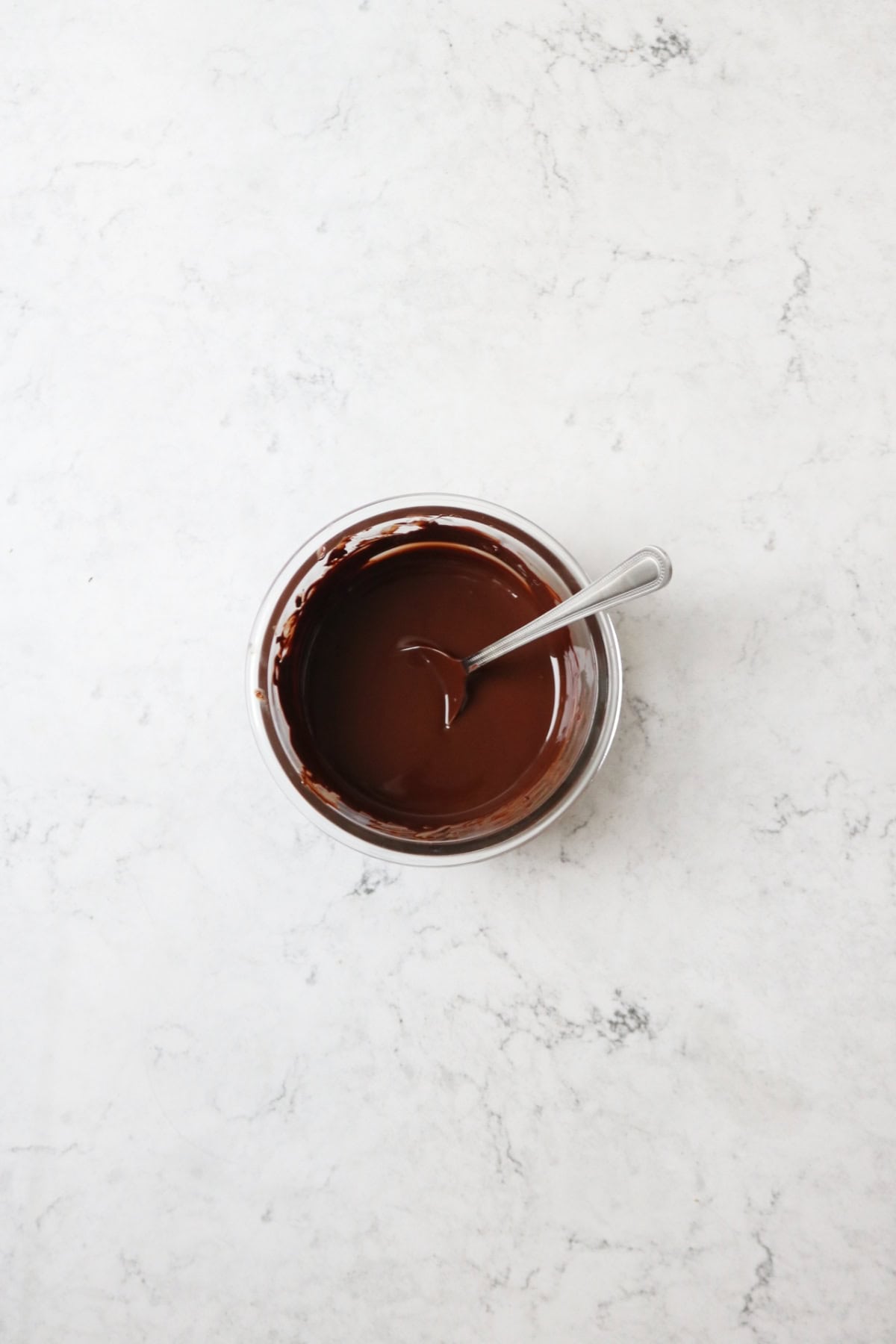 A glass bowl containing melted dark chocolate and a metal spoon